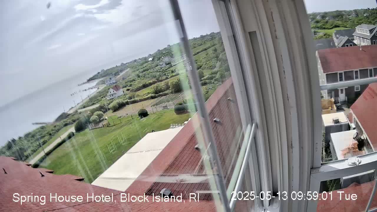 A view from a window shows a partly cloudy day overlooking a coastal town with green lawns, buildings, and a body of water in the distance.