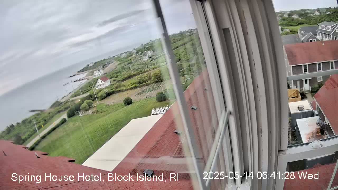 A view from a window shows a cloudy coastal scene with green lawns, buildings, and a body of water under an overcast sky.