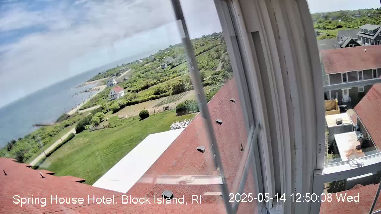 A view from a window shows a partly sunny day overlooking a coastal area with green lawns, buildings, and a body of water in the distance.