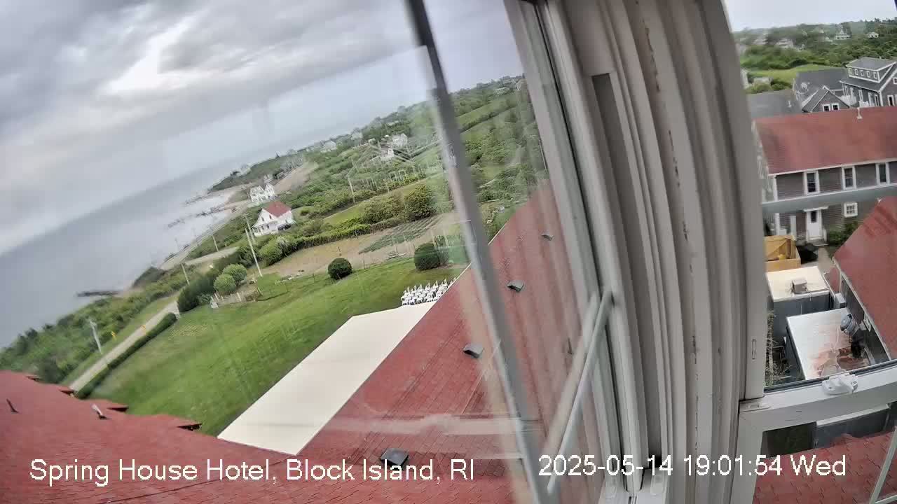 A view from a window shows an overcast day with a coastal scene of green lawns, houses, and a body of water under a cloudy sky.