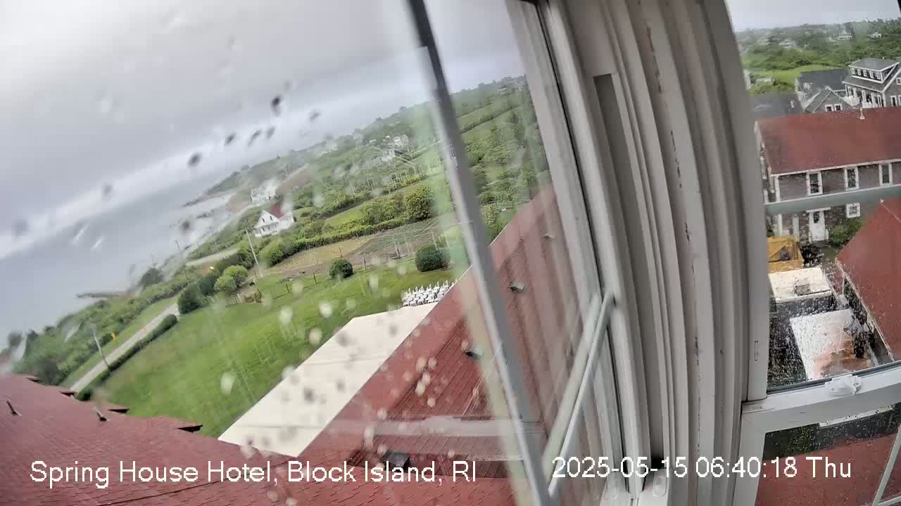 A rain-streaked window reveals a coastal view of houses, grassy areas, and ocean, under a cloudy sky.