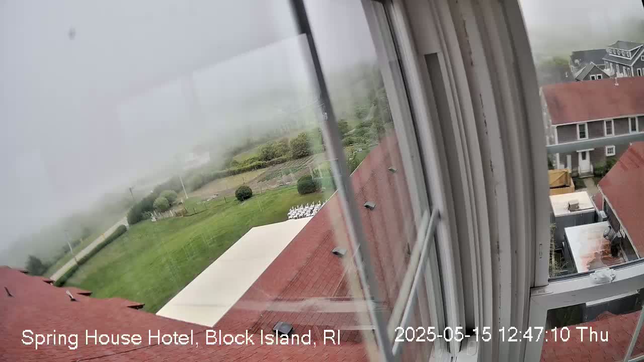 A foggy view from a window shows a red-roofed building, a lawn, and other buildings in the distance.