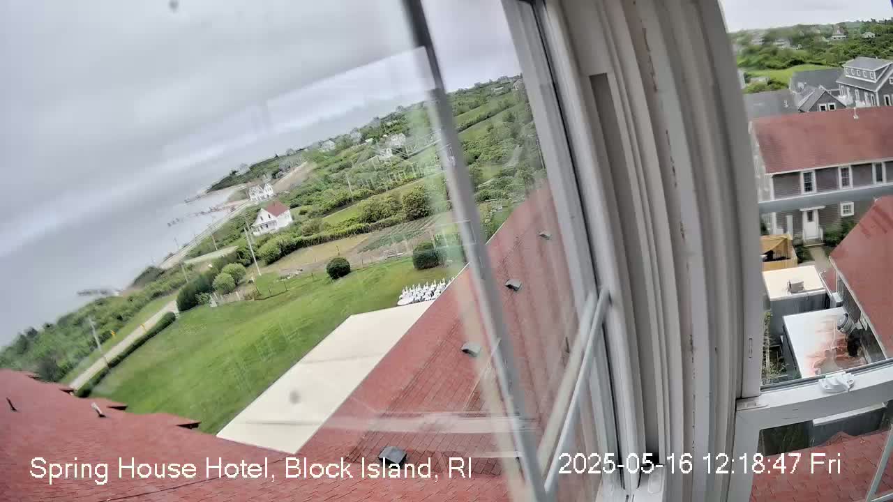 A view from a window shows a cloudy day overlooking a coastal town with houses, grassy areas, and a body of water in the distance.