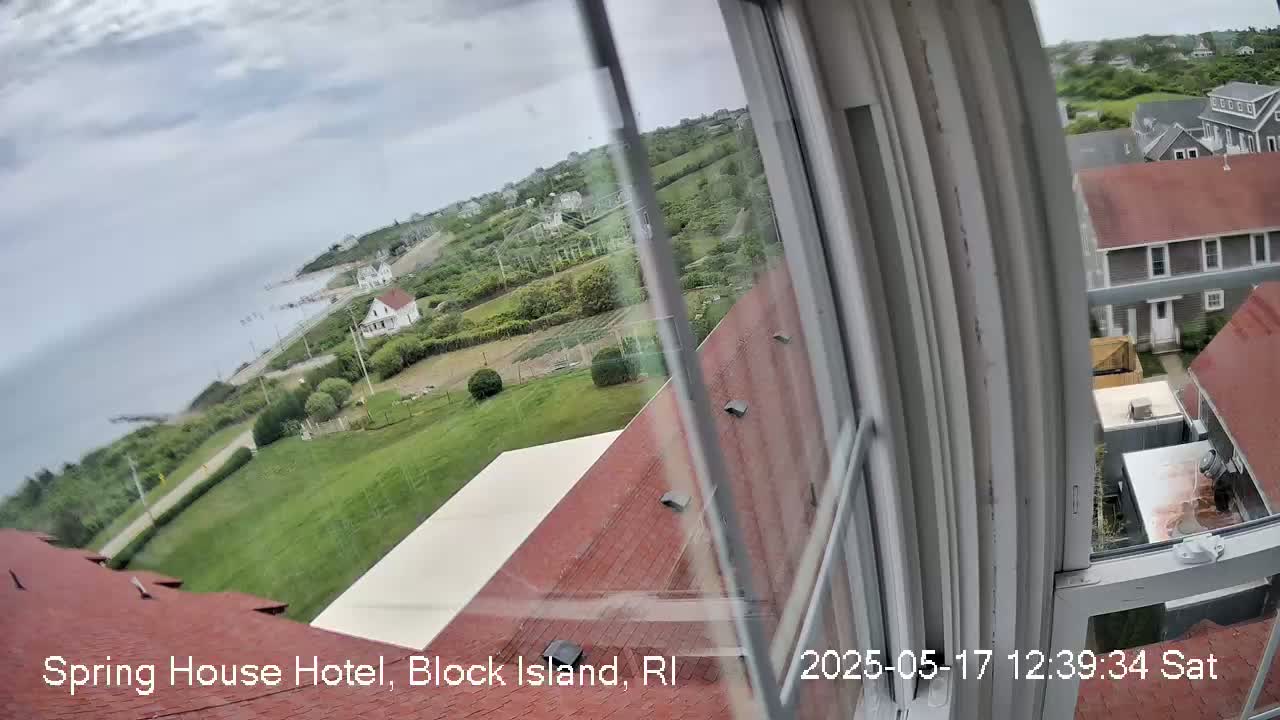 A view from a window shows a coastal scene with overcast skies, green lawns, buildings with red roofs, and a body of water in the distance.