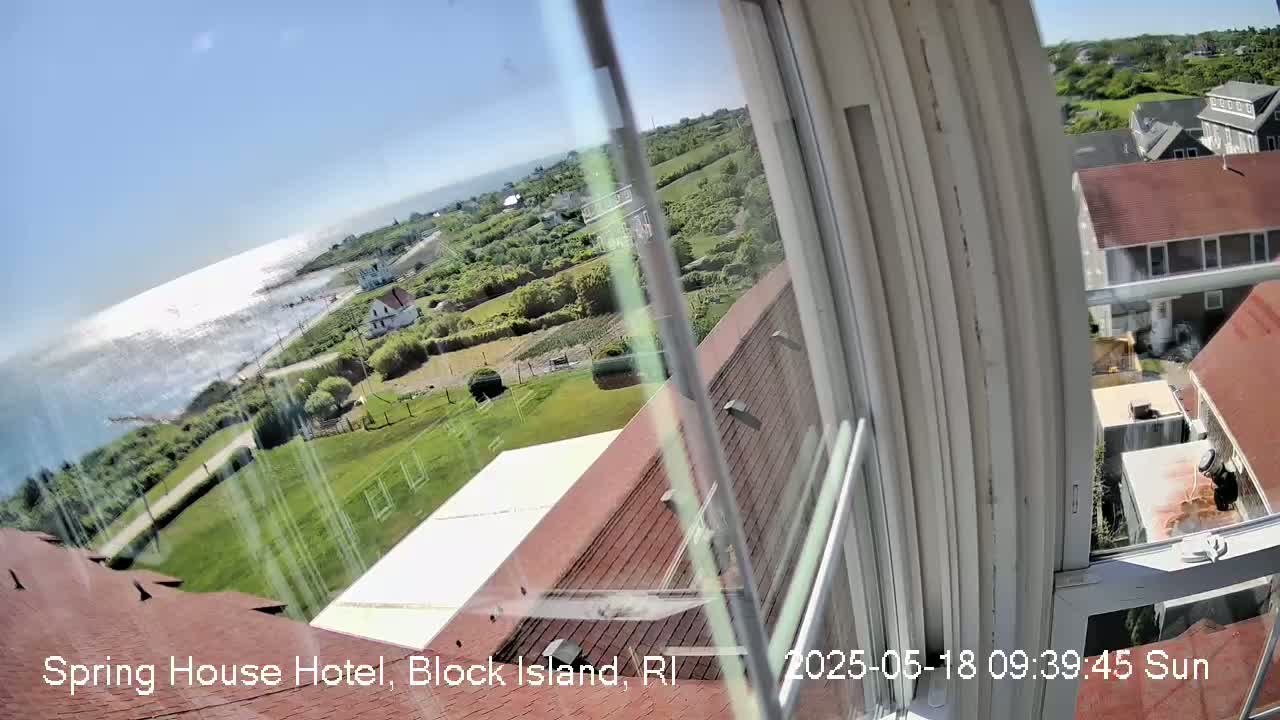 A sunny view of the ocean and coastal buildings is seen from a window, with grassy areas and red rooftops visible.