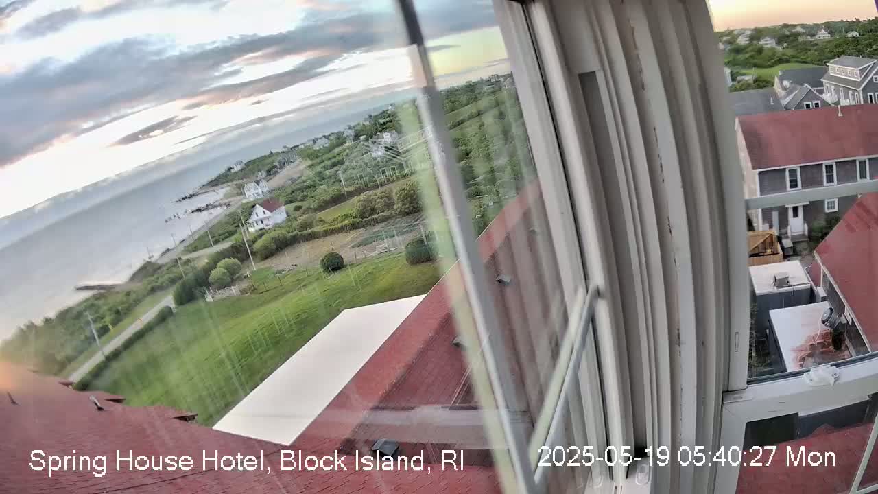 A view from a window shows a partly cloudy sky over a coastal area with buildings, green lawns, and a body of water in the distance.