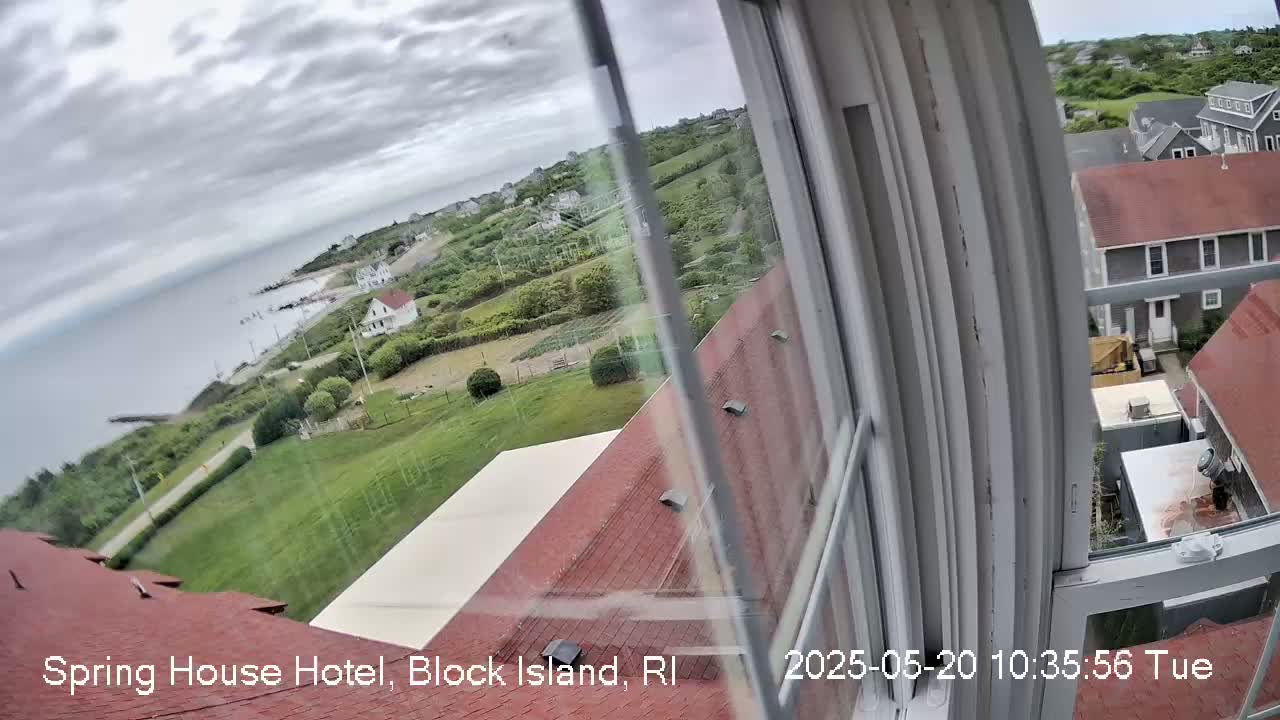A view from a window shows a cloudy coastal scene with green lawns, houses, and a body of water.
