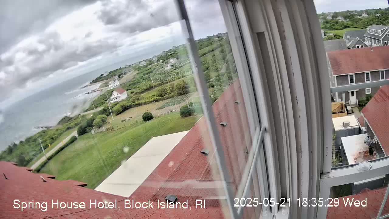 A view from a window shows a cloudy day overlooking a coastal area with houses, green lawns, and a body of water.