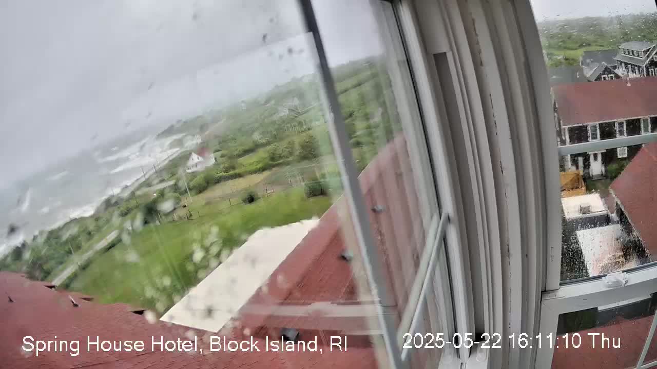 A rain-streaked window shows a view of a stormy ocean with crashing waves, green coastal land, and buildings under a gray sky.
