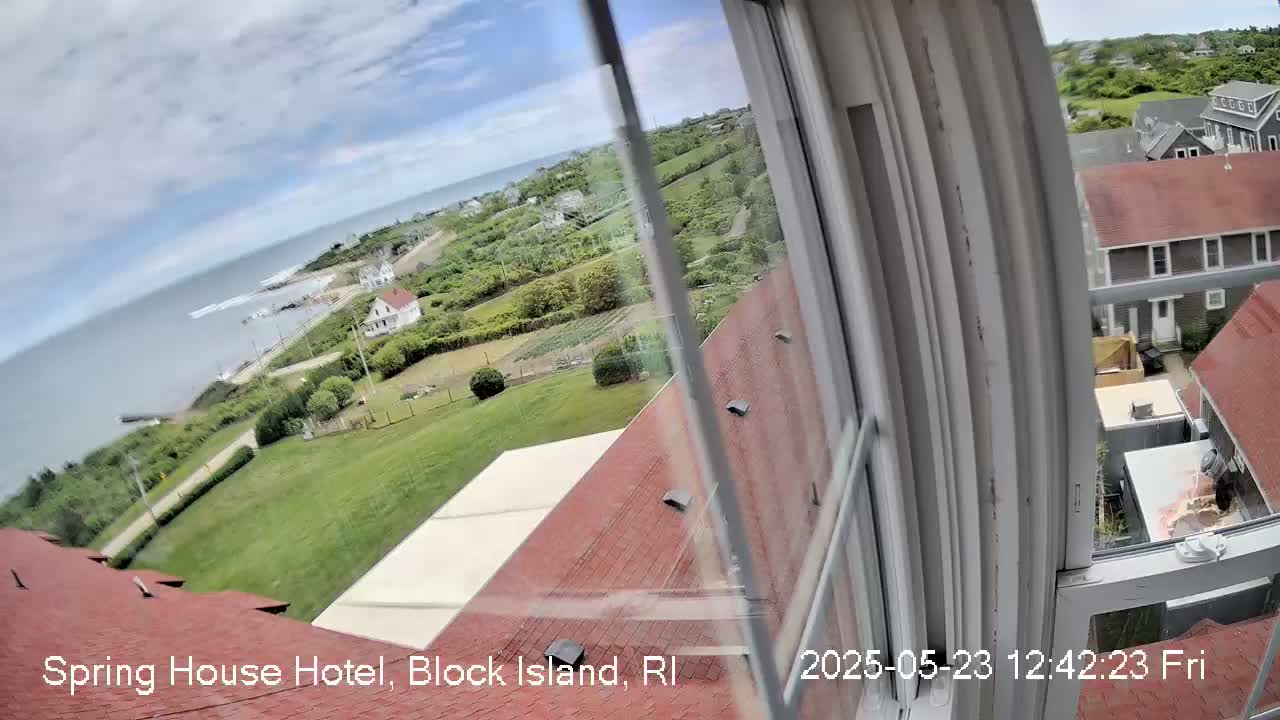 A partly cloudy day overlooks a coastal scene of grassy fields, houses, and a calm ocean from a window with a red roof in the foreground.