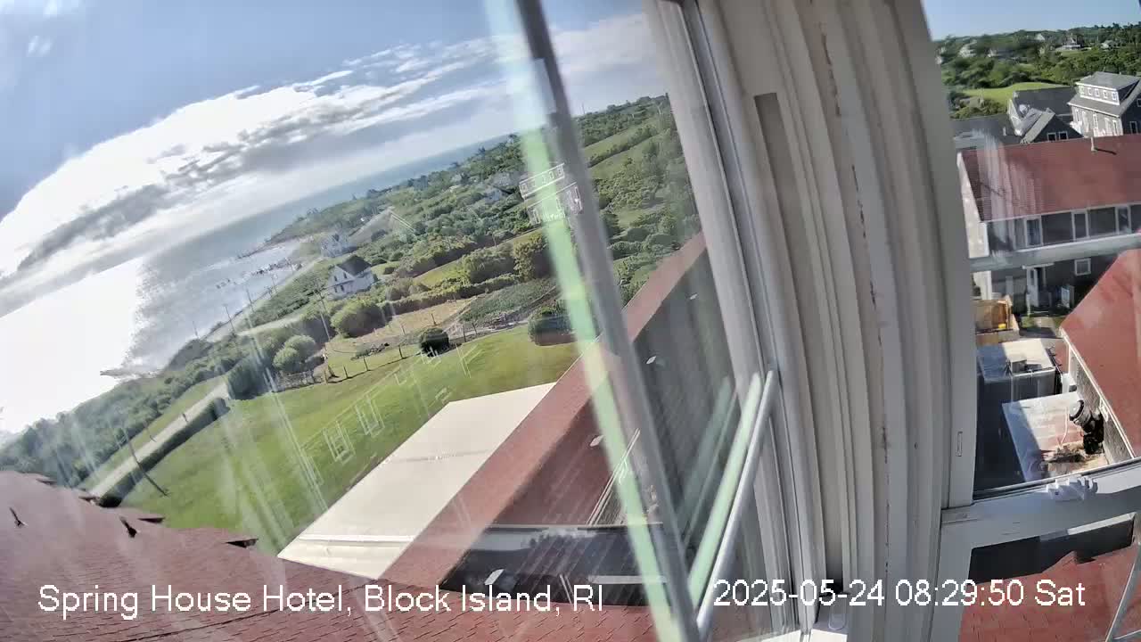 A view from a window shows a partly cloudy sky, ocean, green lawns, buildings, and a partially visible awning, all seen from an elevated position.