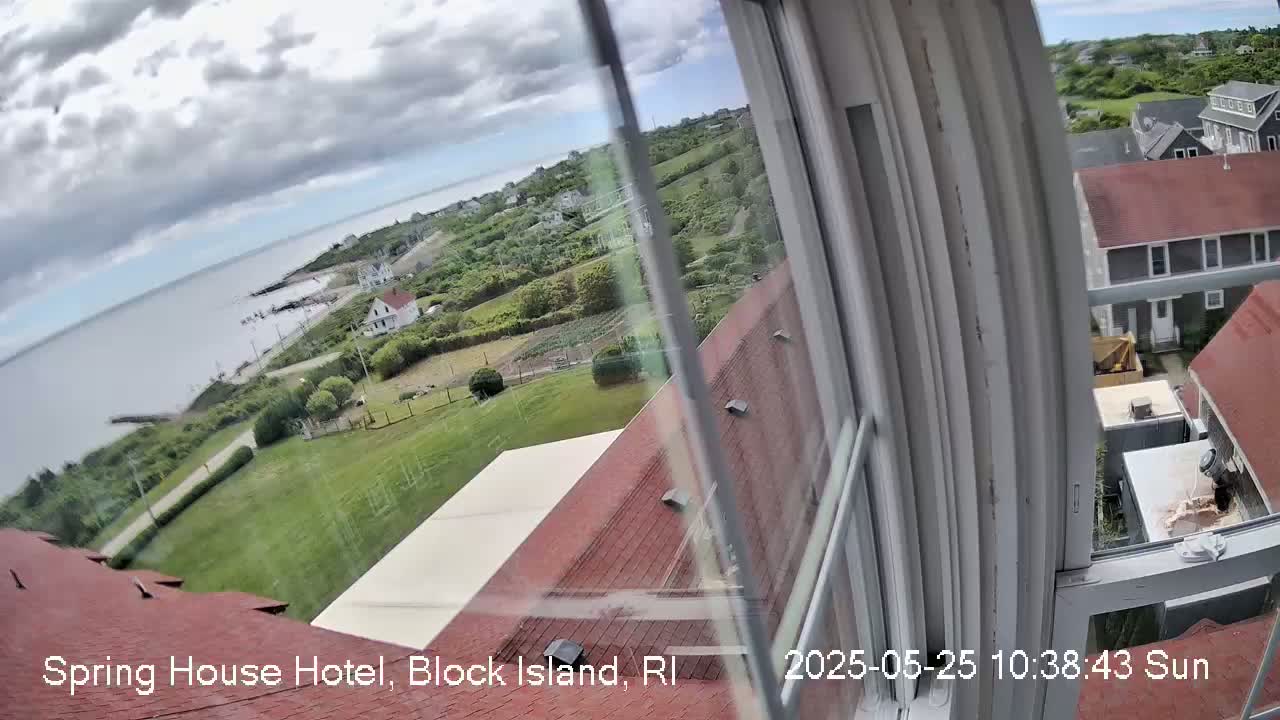A partly cloudy view from a window shows a coastal scene with green lawns, houses, and a body of water under a mostly cloudy sky.