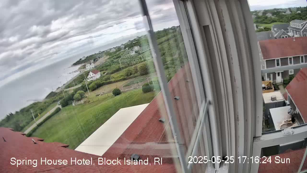 A view from a window shows a cloudy day overlooking a coastal town with houses, green spaces, and a body of water in the distance.