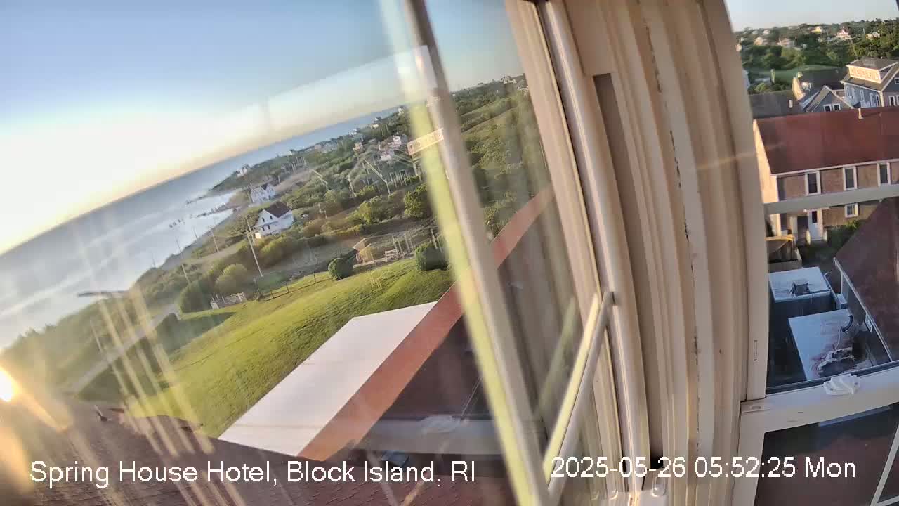 A view through a window shows a calm ocean, grassy landscape, houses, and a clear, sunny sky.
