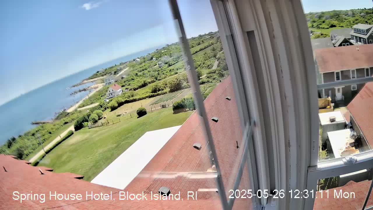 A view from a window shows a sunny day overlooking a coastal area with buildings, green lawns, and a body of water.