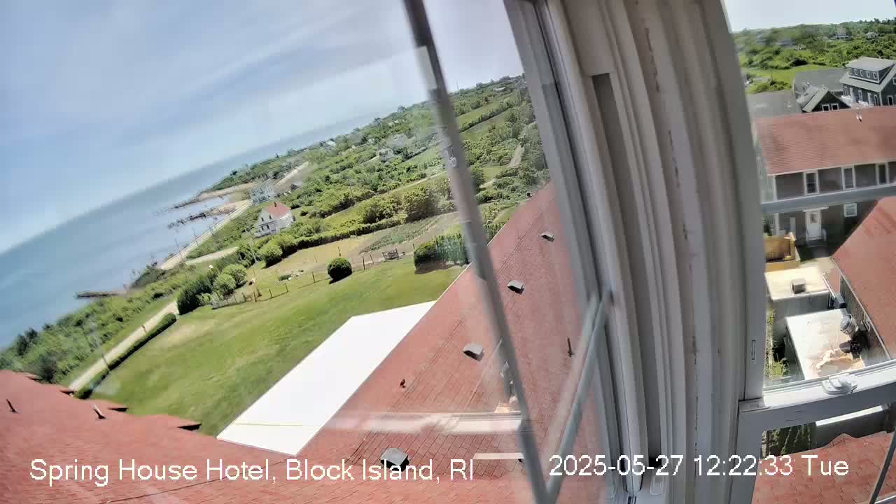 A view from a window shows a coastal scene on a sunny day, with green lawns, buildings, and ocean visible beyond a road.