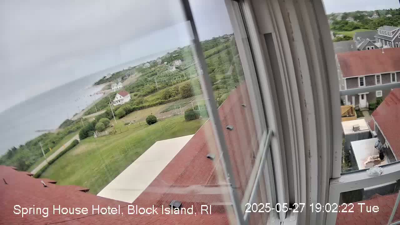 A view from a window shows a cloudy day overlooking a coastline with houses and green lawns, partially obscured by the window frame.