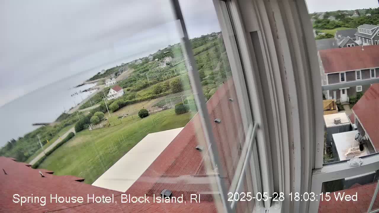A view from a window shows a cloudy day overlooking a coastal town with green lawns, houses, and the ocean in the distance.
