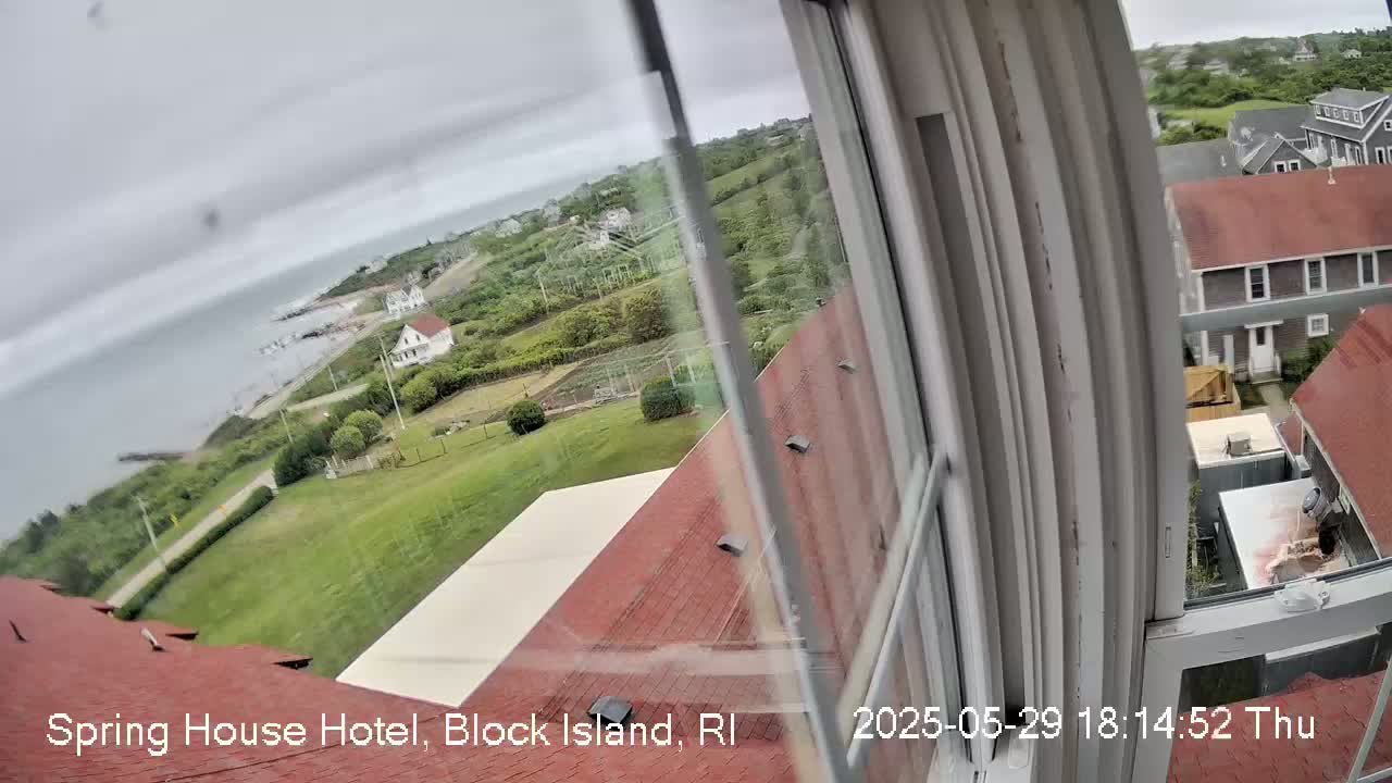 A view from a window shows a cloudy day overlooking a coastal area with houses, green lawns, and a body of water.