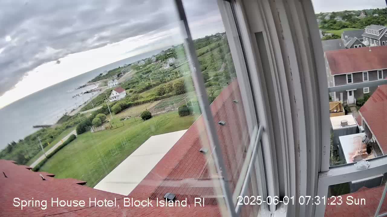 A view from a window shows a cloudy day overlooking a coastal area with green lawns, buildings, and a body of water.