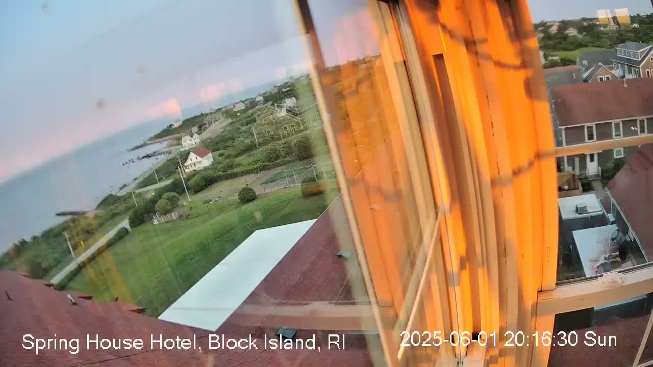 A view from a window shows a sunset over a calm ocean, coastal houses, and green lawns.