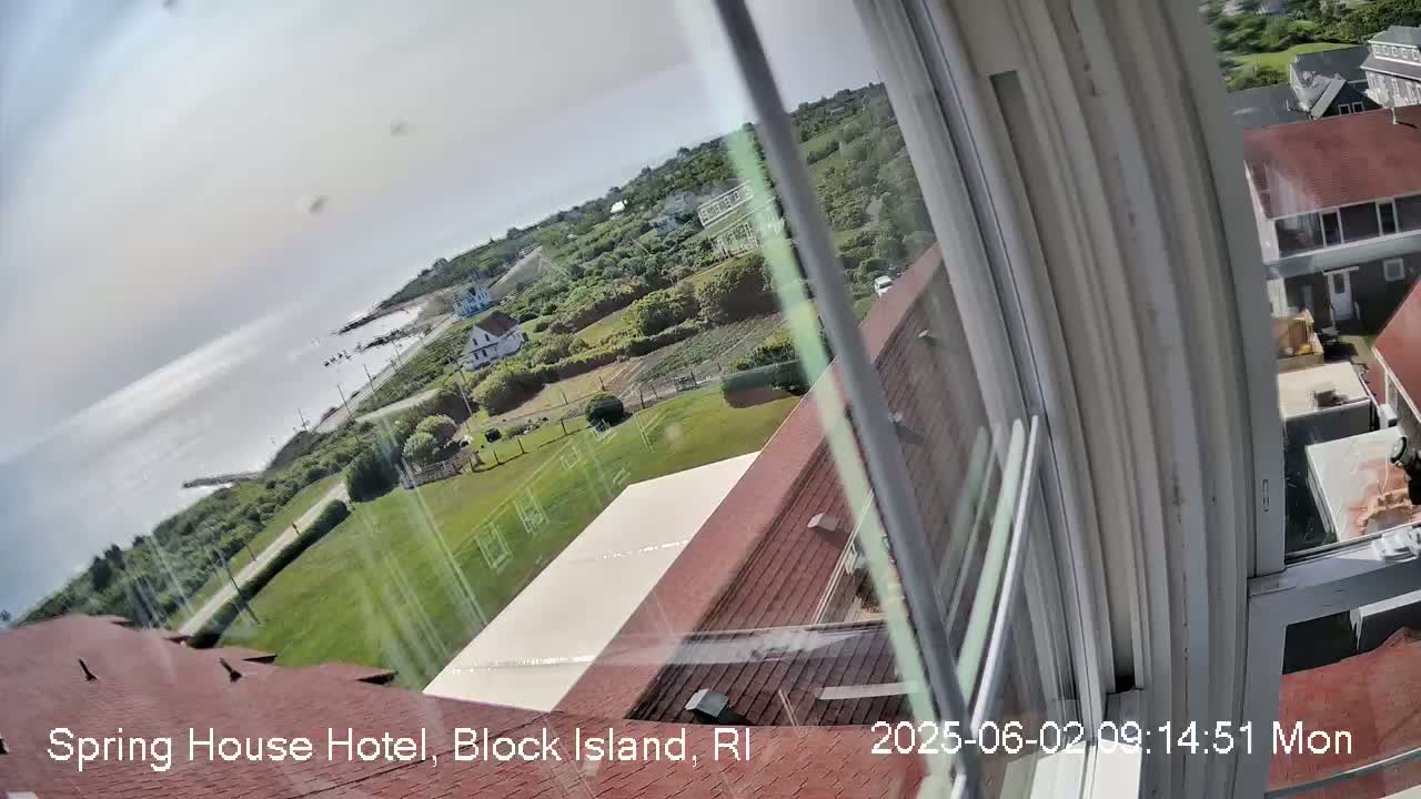 A view from a window shows a partly cloudy day overlooking a coastal area with green lawns, buildings, and a calm ocean.