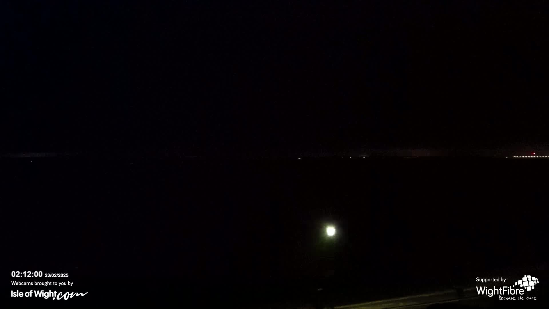 A nighttime view of a dark body of water with faint lights on the horizon and a single lamp visible in the foreground.