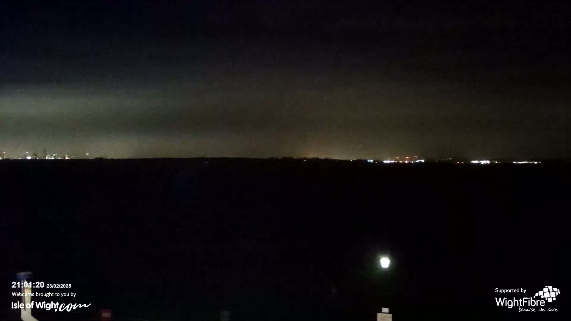 A dark, nighttime landscape shows a distant, faintly lit cityscape under a cloudy sky.
