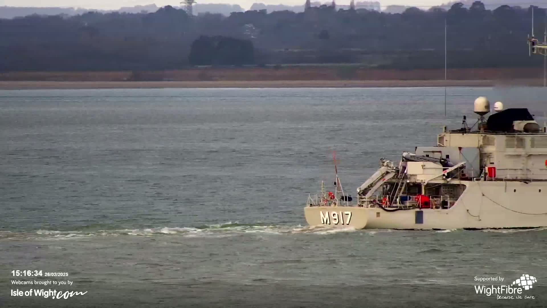 A beige ship numbered M917 sails on a choppy gray body of water with a distant shoreline visible.
