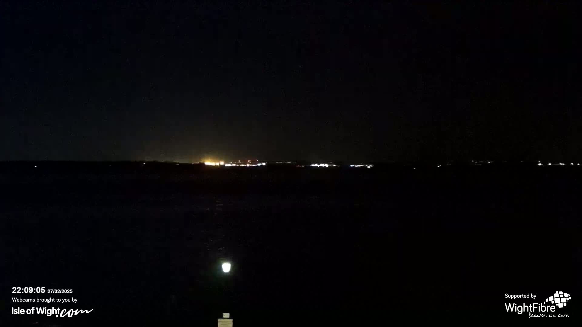 A dark, nighttime scene shows a distant line of lights across a body of water under a clear sky.