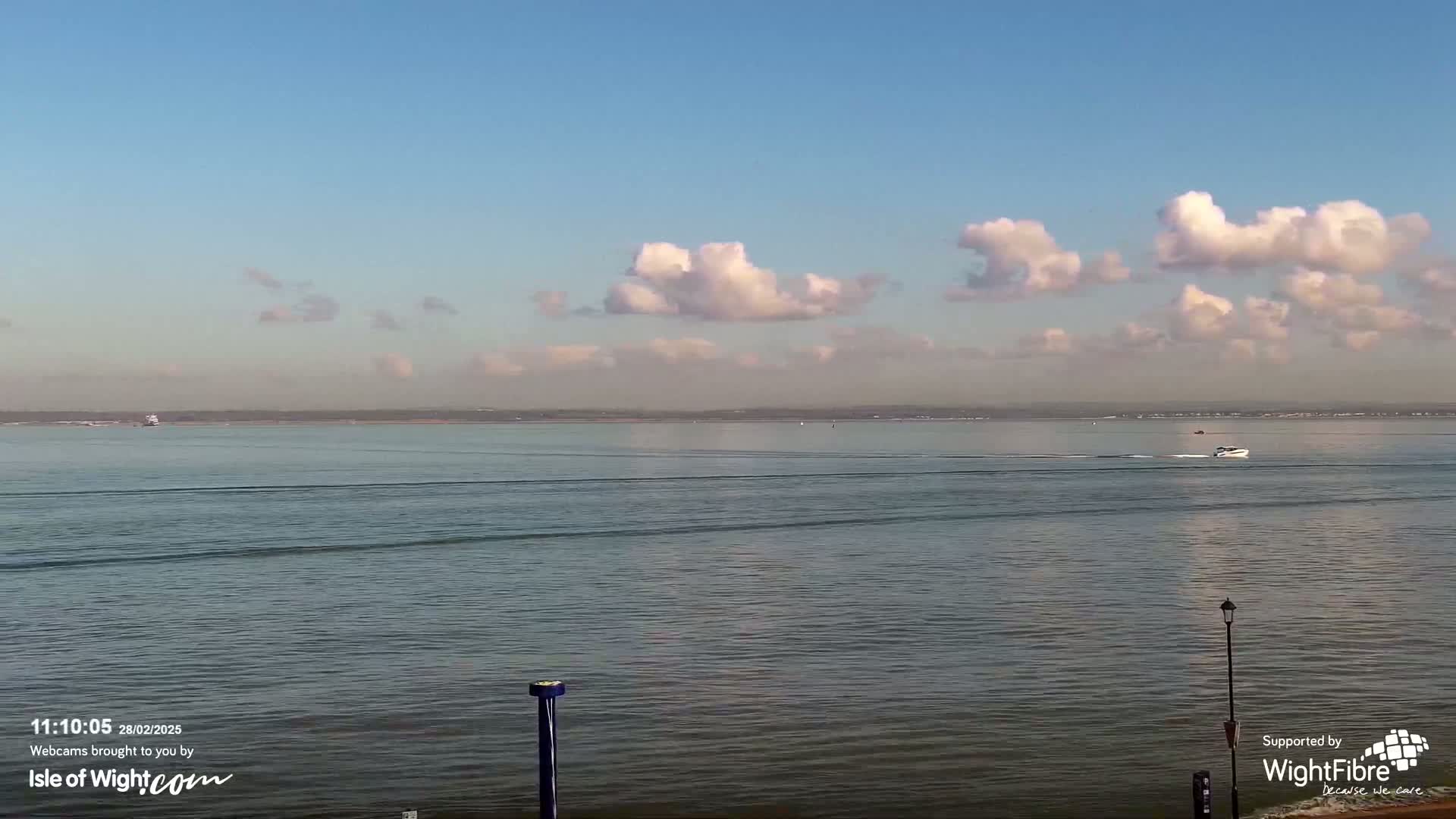 A calm body of water stretches to a distant shoreline under a mostly clear sky with scattered puffy clouds.