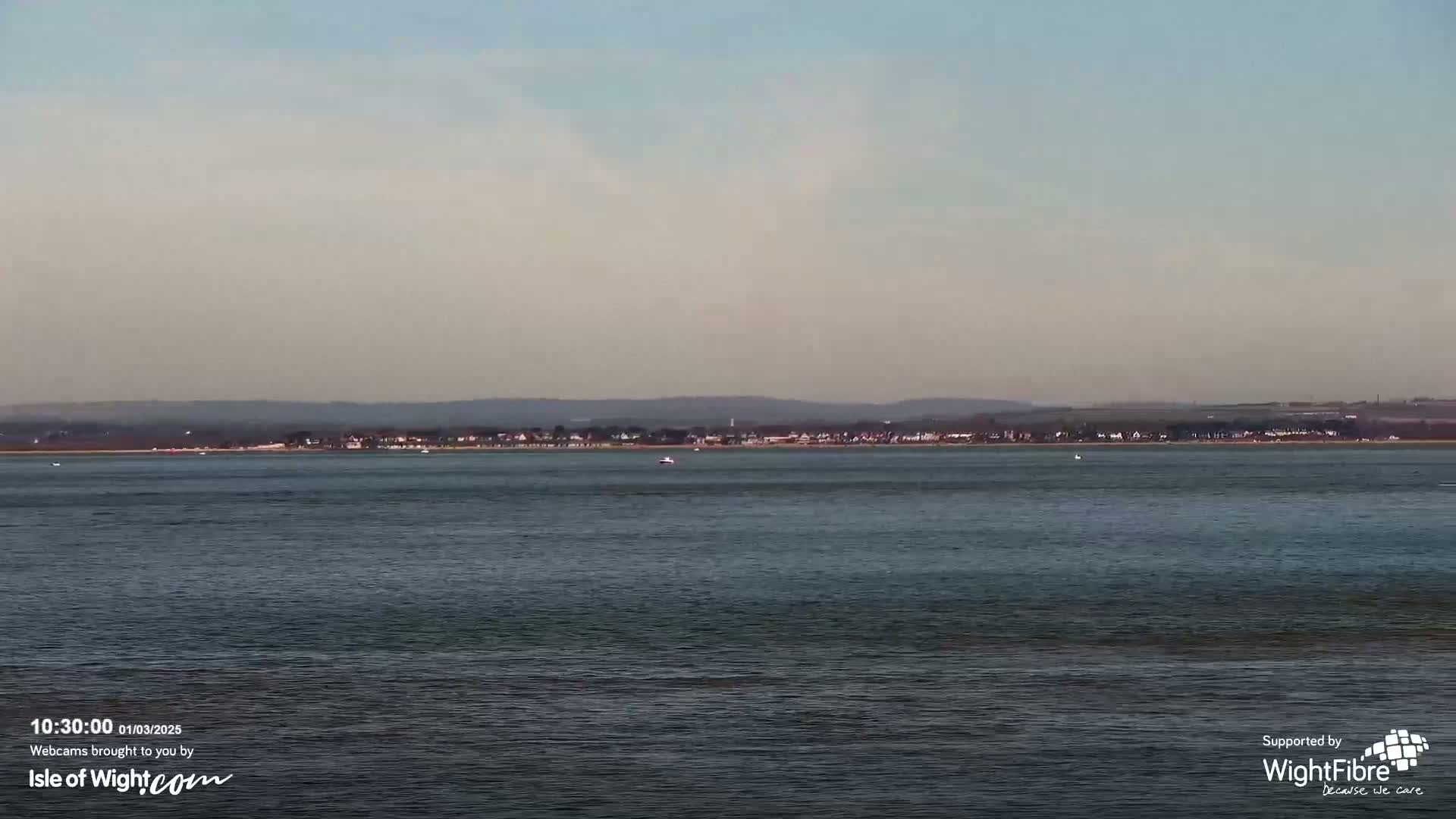 A calm body of water shows a distant shoreline with buildings under a hazy sky.