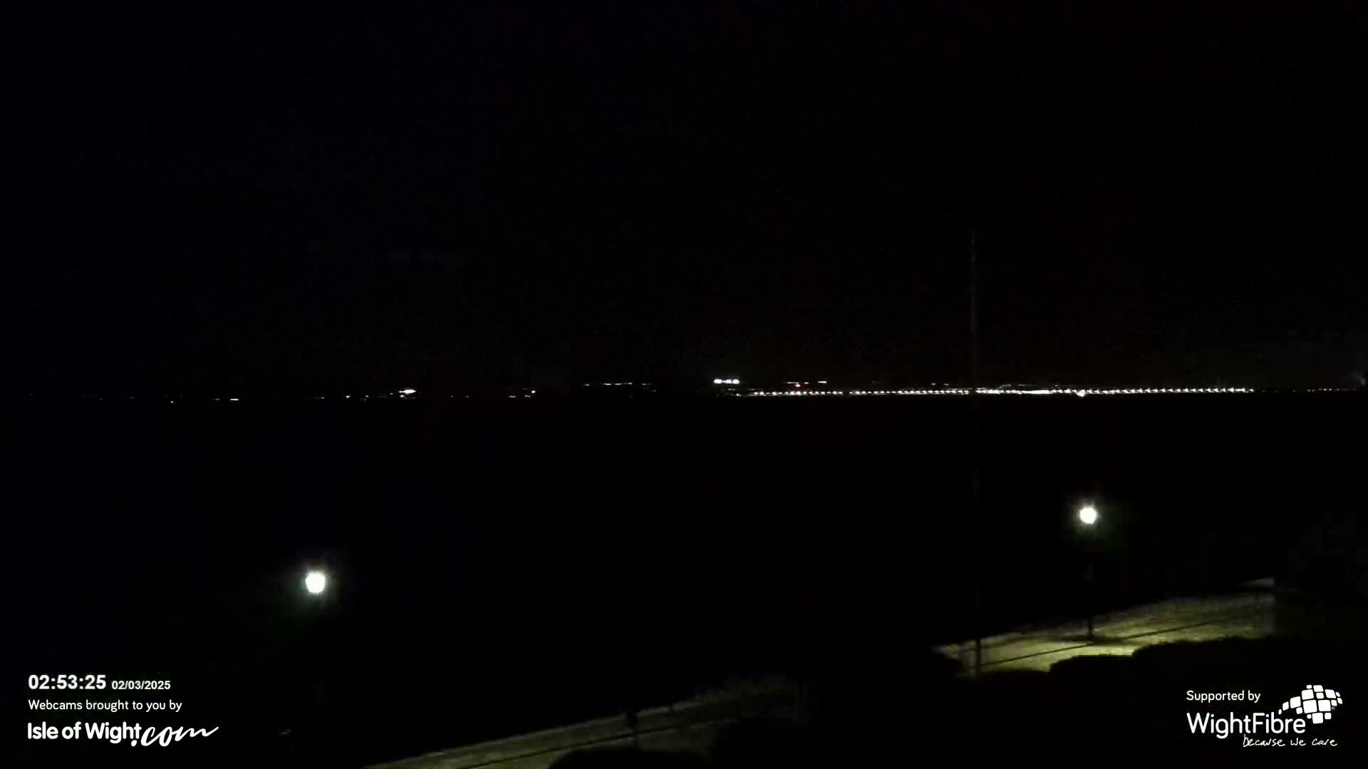 A nighttime long shot shows a distant line of city lights across a dark body of water, with a few illuminated streetlights visible in the foreground.