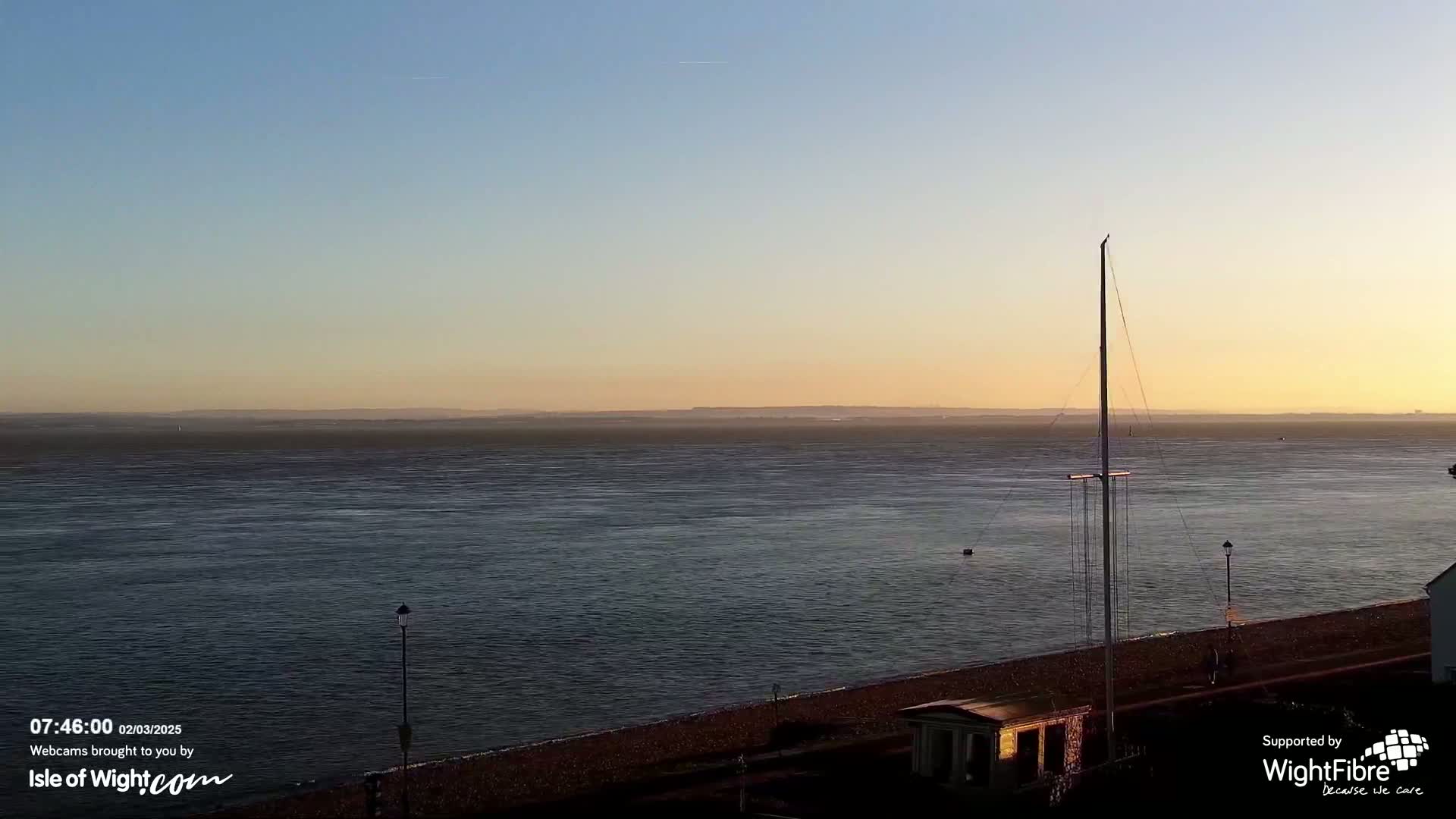 A calm body of water stretches to a distant shoreline under a clear, sunset sky, with a tall mast and small structures visible near the water's edge.