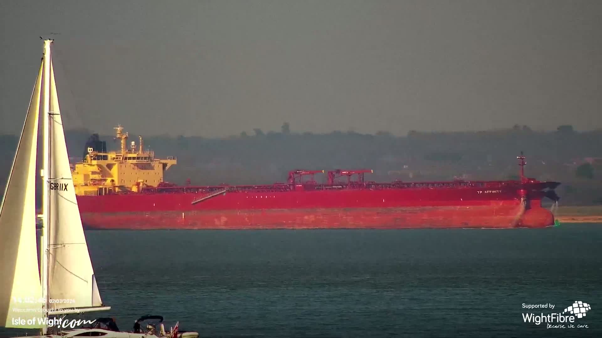 A large red cargo ship is visible in the distance on calm water, with a sailboat in the foreground.
