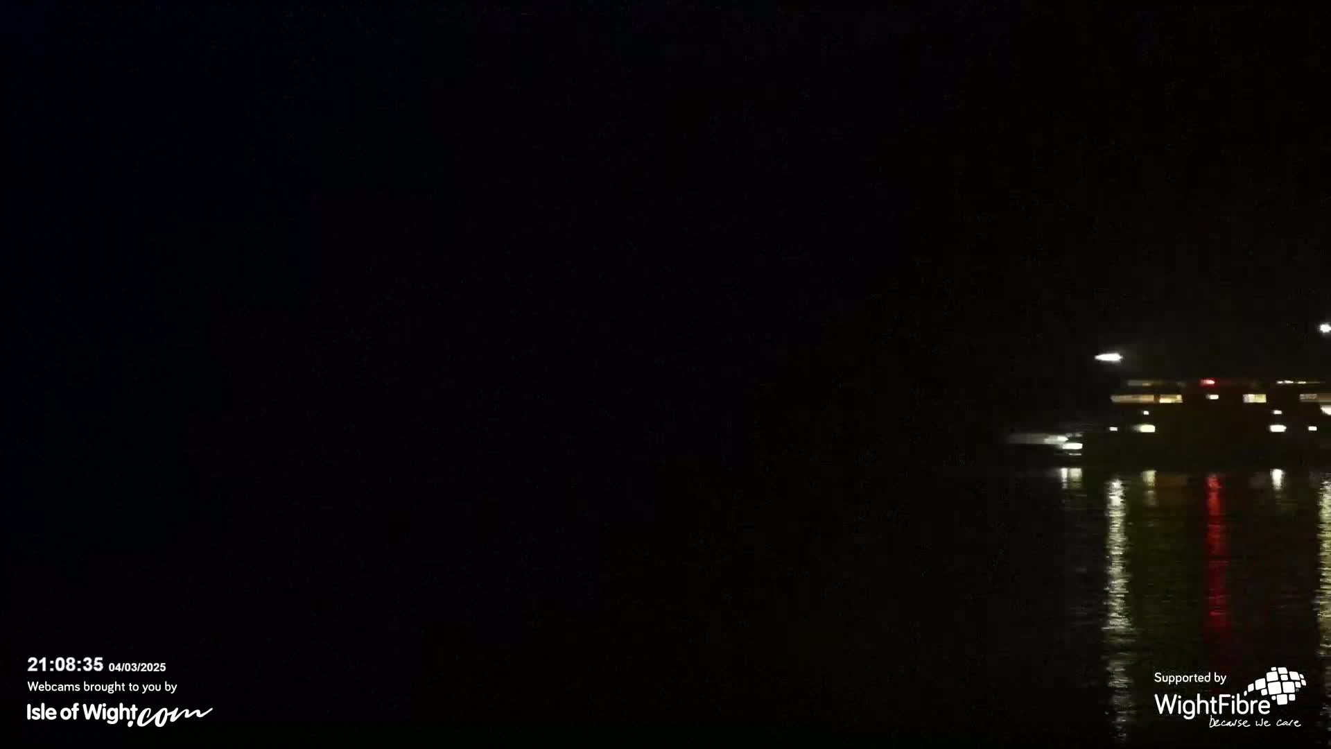 A dark, nighttime scene shows a lit-up building reflected in calm water.