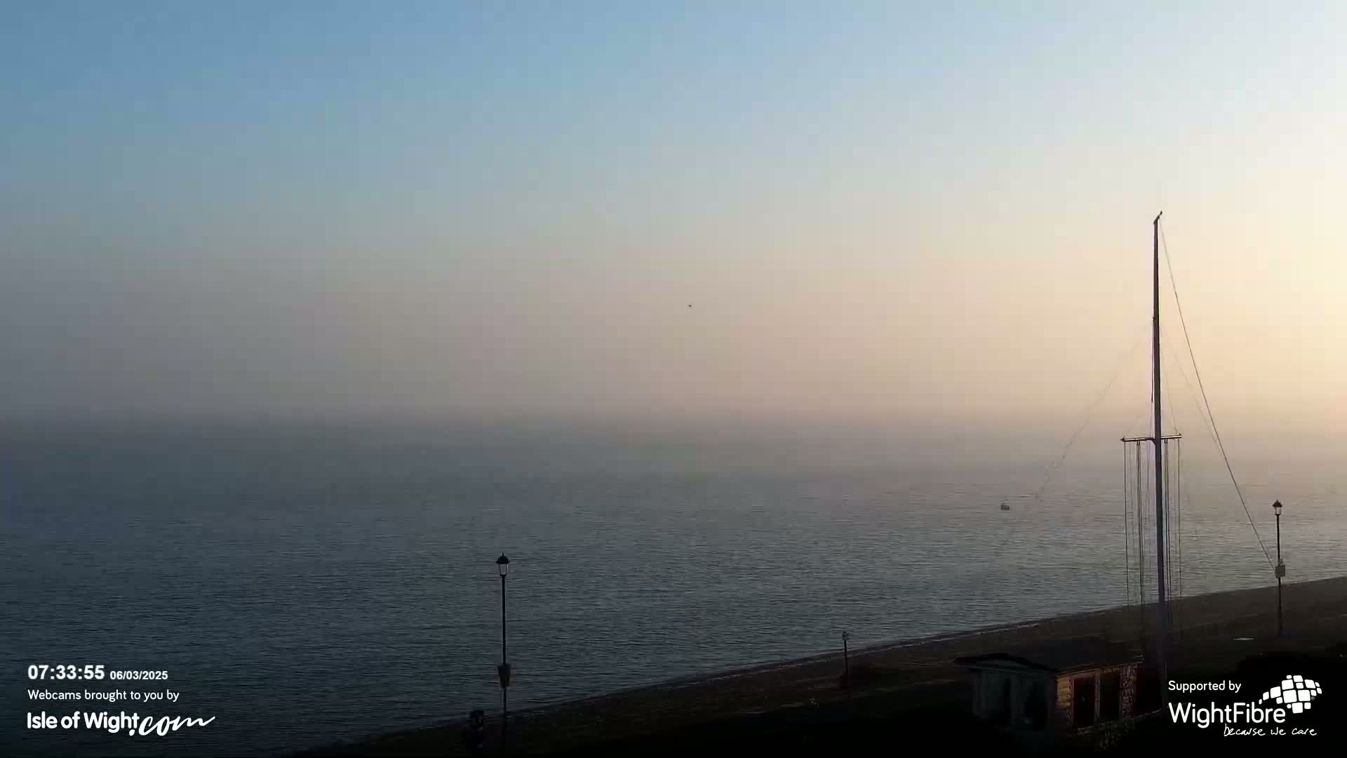 A hazy sunrise over a calm sea with a tall mast and small boats in the distance, viewed from a shoreline with lampposts.