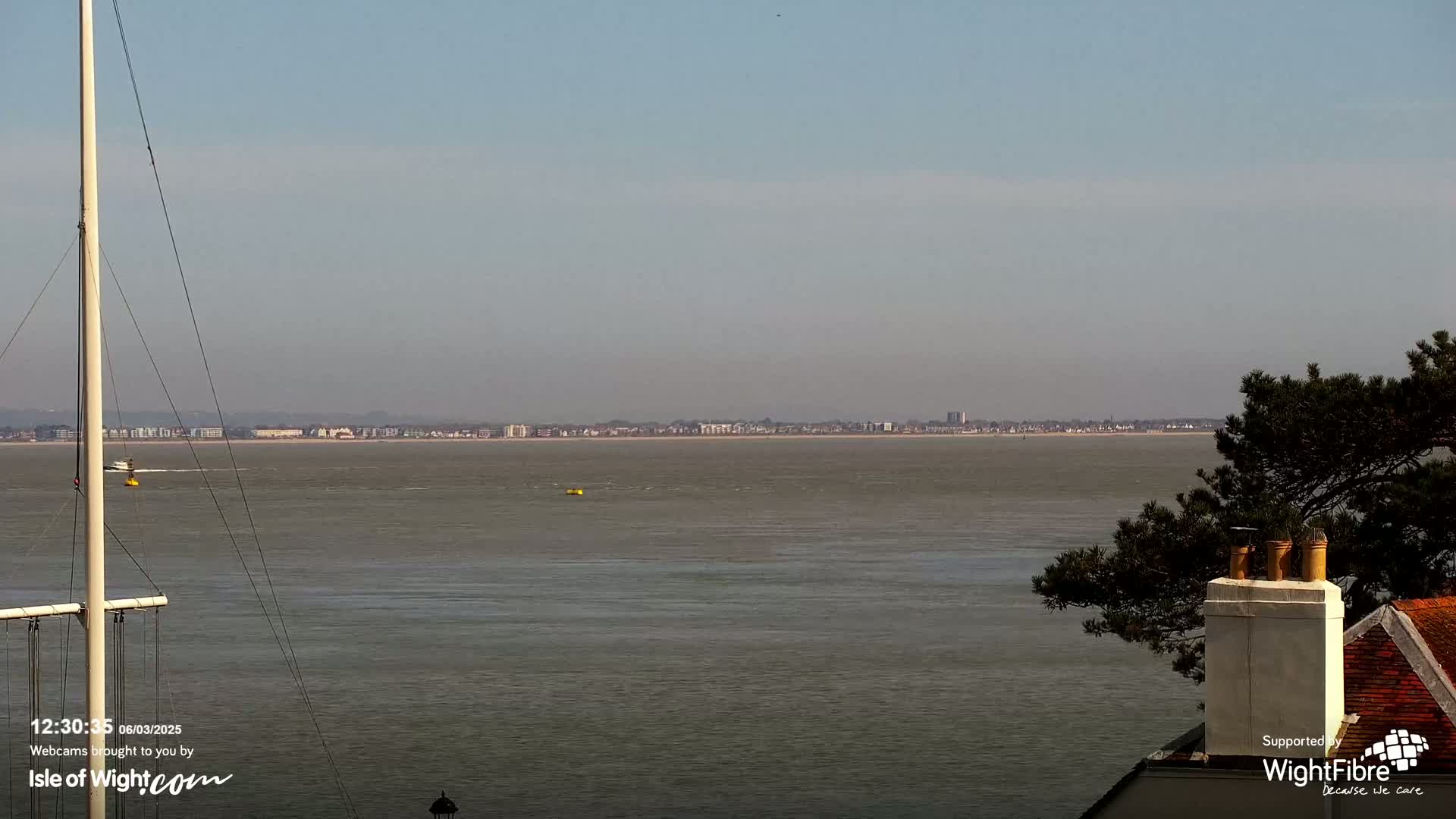 A calm, wide body of water stretches to a distant shoreline of buildings under a clear sky, with a sailboat mast and a portion of a building visible in the foreground.