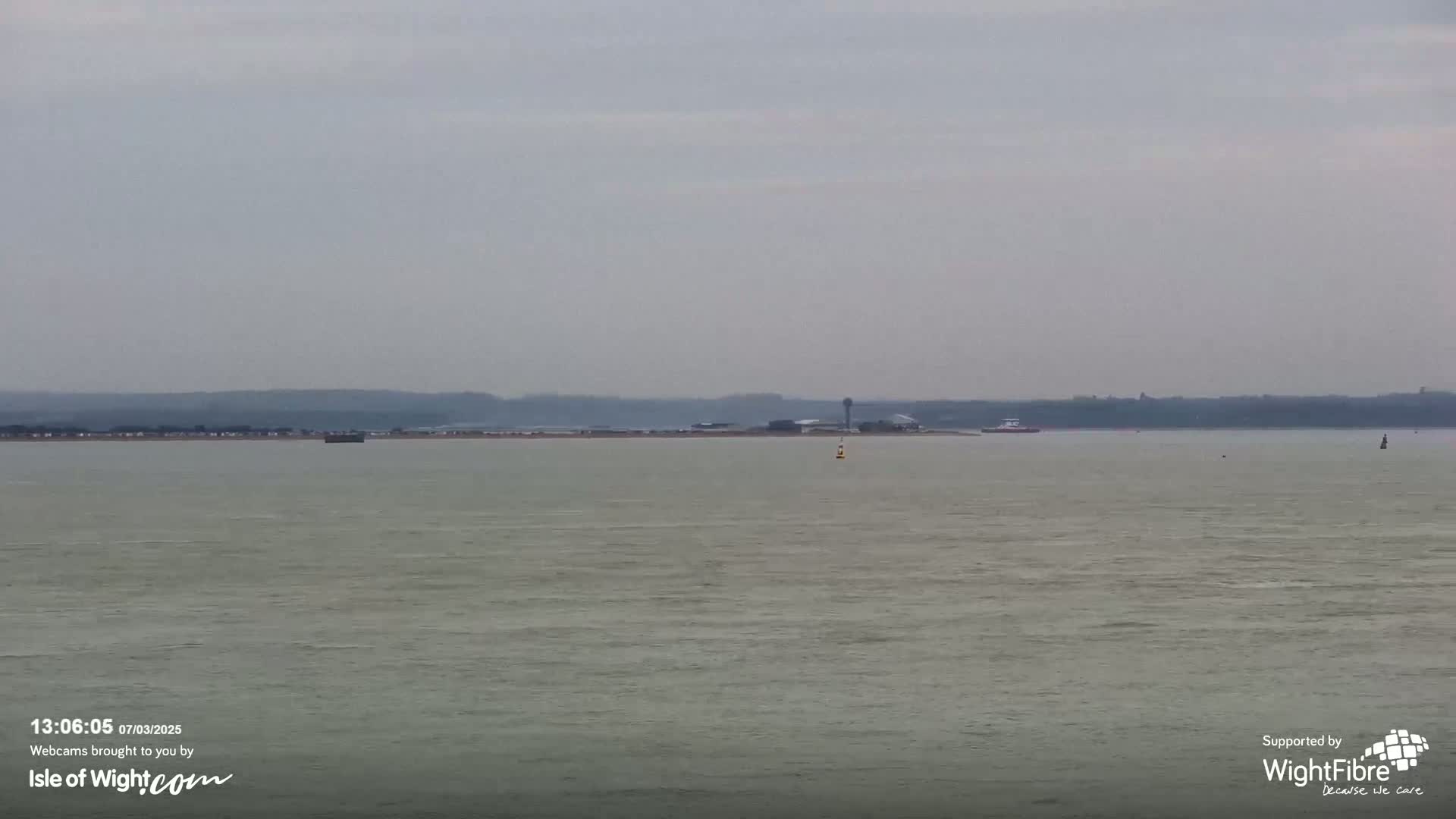 A calm, overcast day reveals a body of water with a distant shoreline featuring a low-lying landmass and some structures.