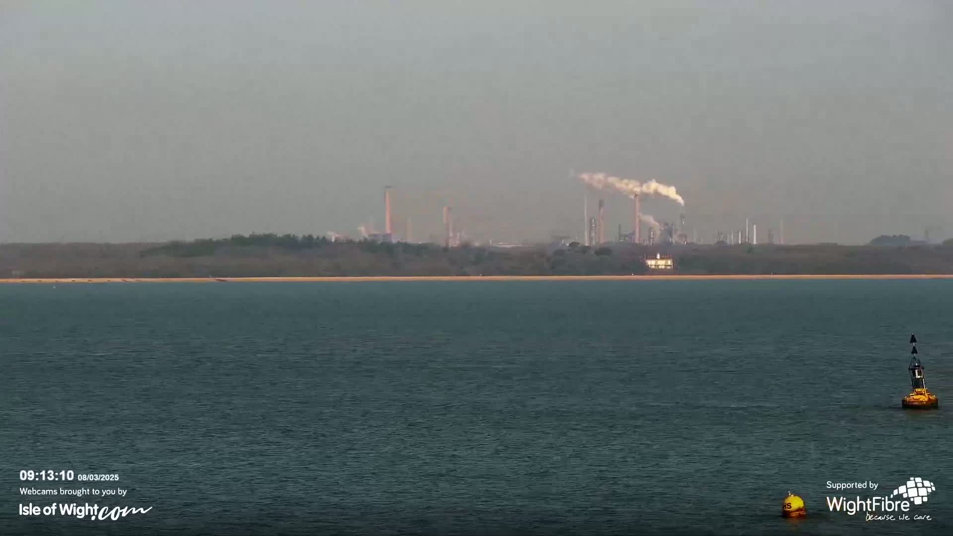 A hazy view shows a body of water with a distant industrial plant emitting smoke, behind a low-lying shoreline.