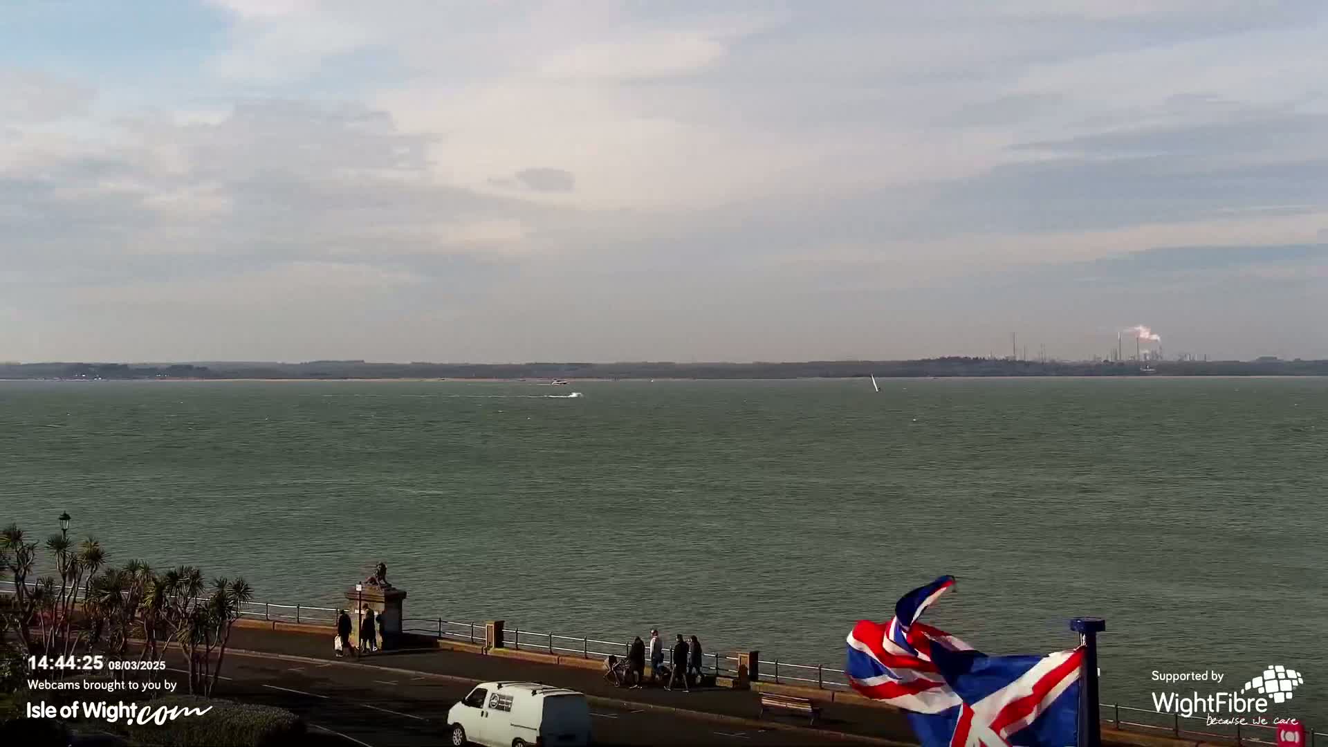 A partly cloudy day shows a wide expanse of calm, greenish-grey water with a distant industrial area visible,  a shoreline with a few people and a van, and a British flag waving in the breeze.