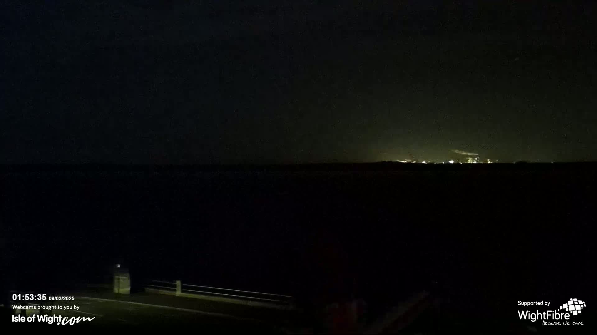 A dark, nighttime scene shows a body of water with faintly lit land in the distance under a clear, dark sky.