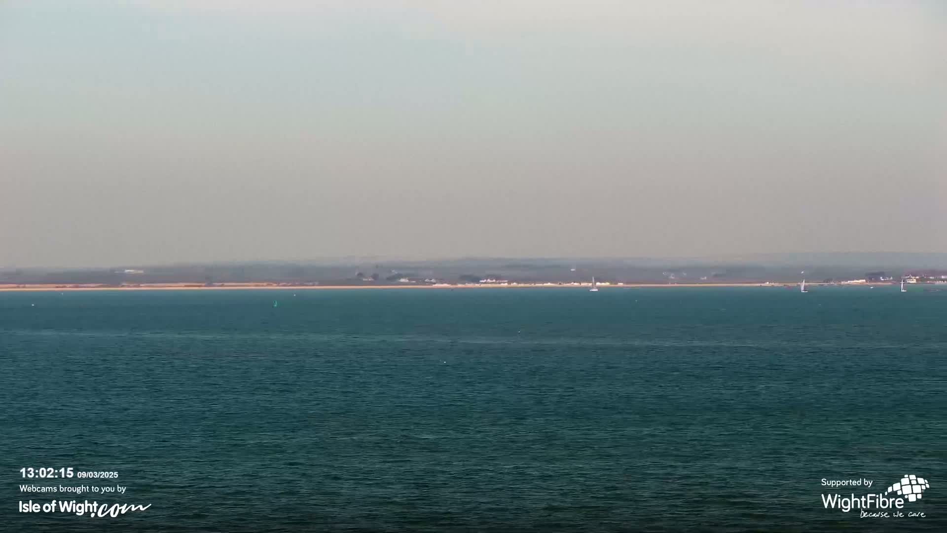 A hazy, distant shoreline with a few small boats is visible across a calm, dark teal sea.