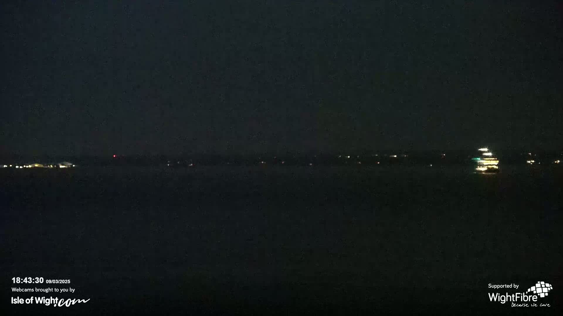 A dark nighttime view of a body of water with a few distant lights on the far shore and a lit-up vessel on the water.
