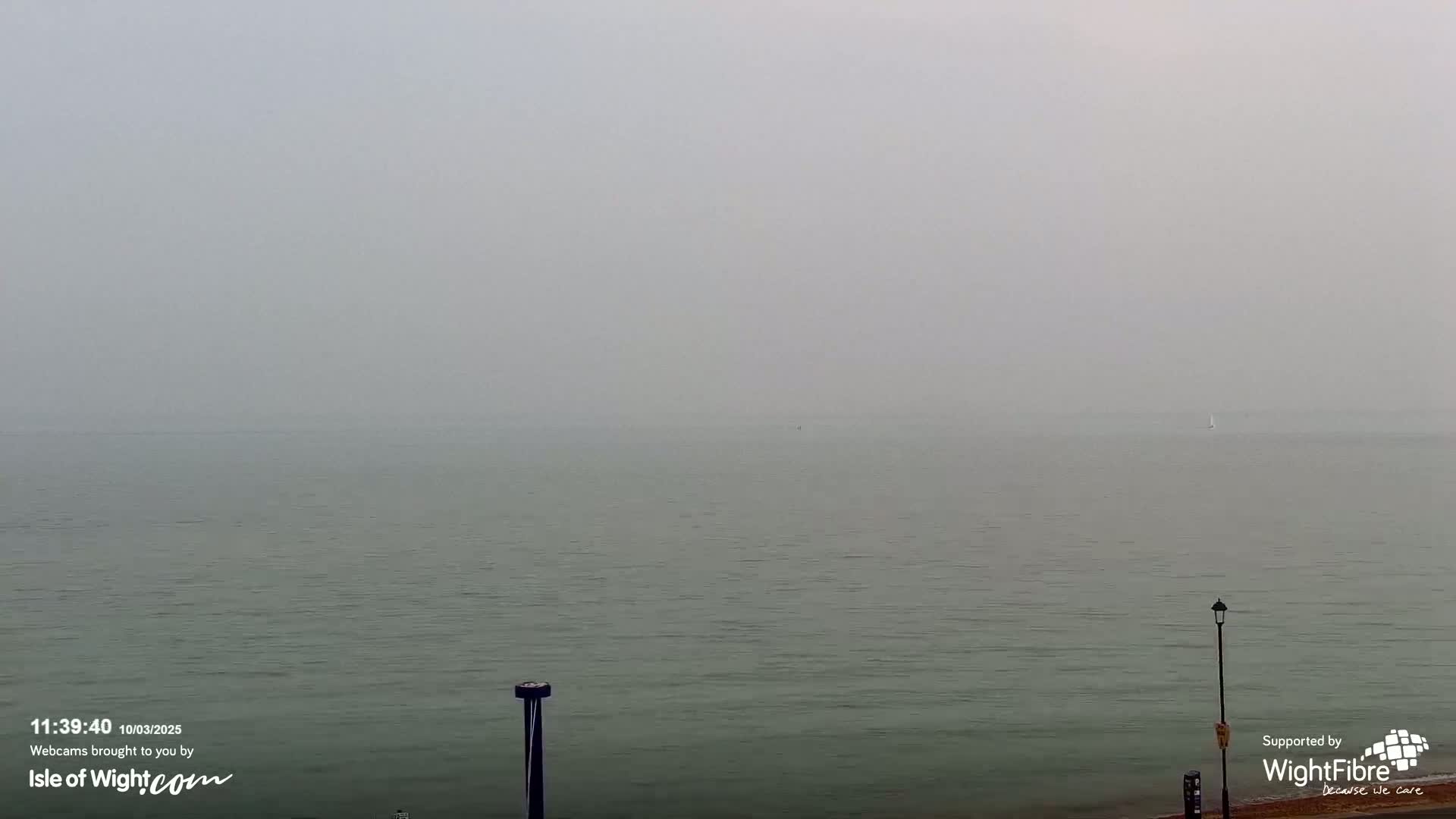 A calm, foggy sea is visible from a shoreline, with a lone sailboat in the distance and a lamppost on the right.