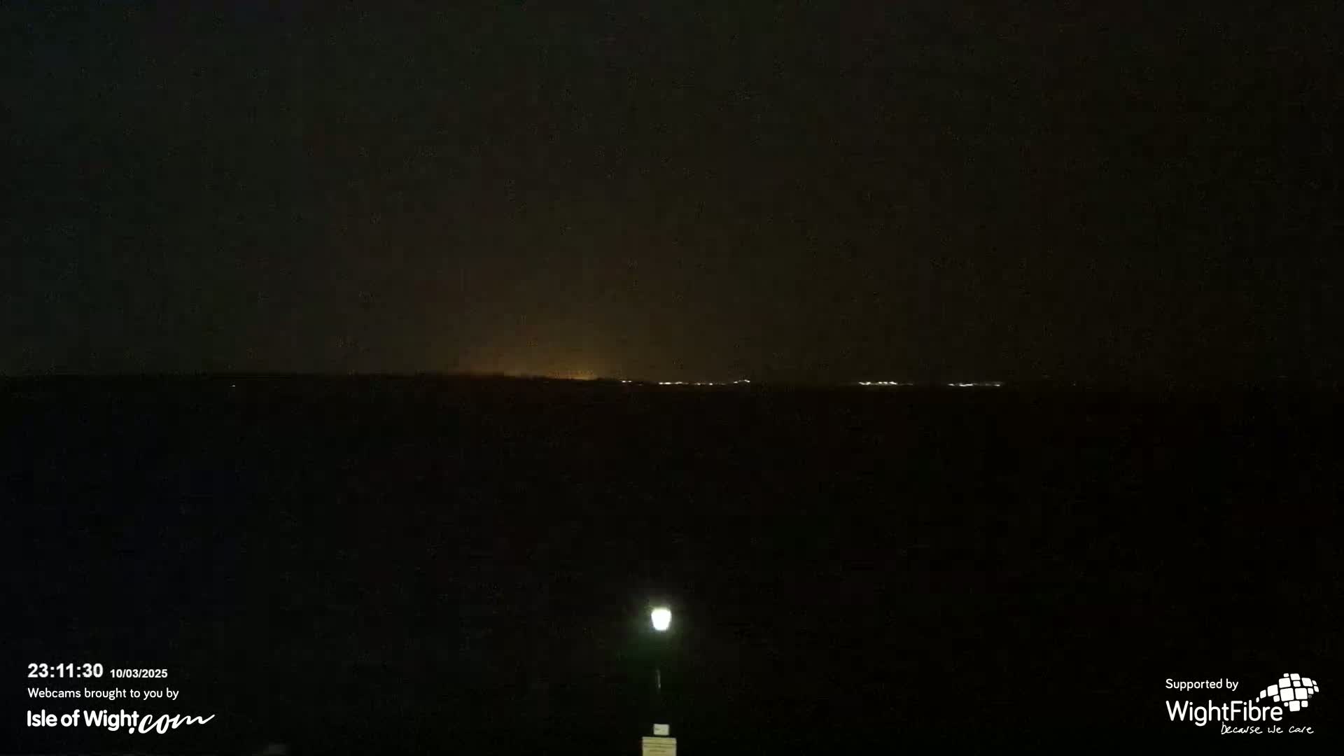 A nighttime, dark scene shows a distant horizon line with faint lights, and a single illuminated lamppost in the foreground.