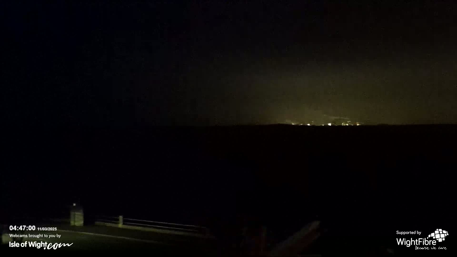 A nighttime, dark outdoor scene shows a distant, faintly lit town or city skyline under a hazy sky.