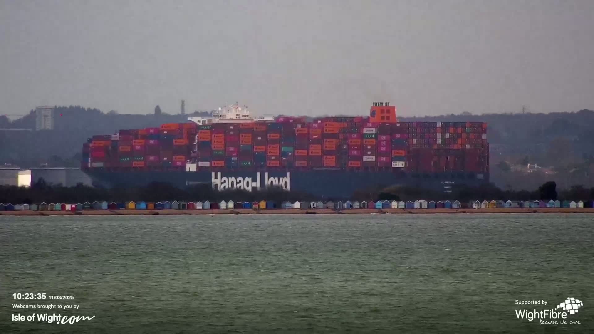 A large container ship, bearing the Hapag-Lloyd logo, is anchored in calm water off a shoreline with a row of colorful beach huts, under an overcast sky.