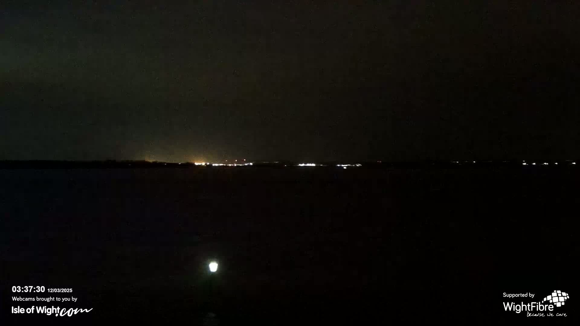 A dark, nighttime scene shows a distant, faintly lit shoreline across a body of water.