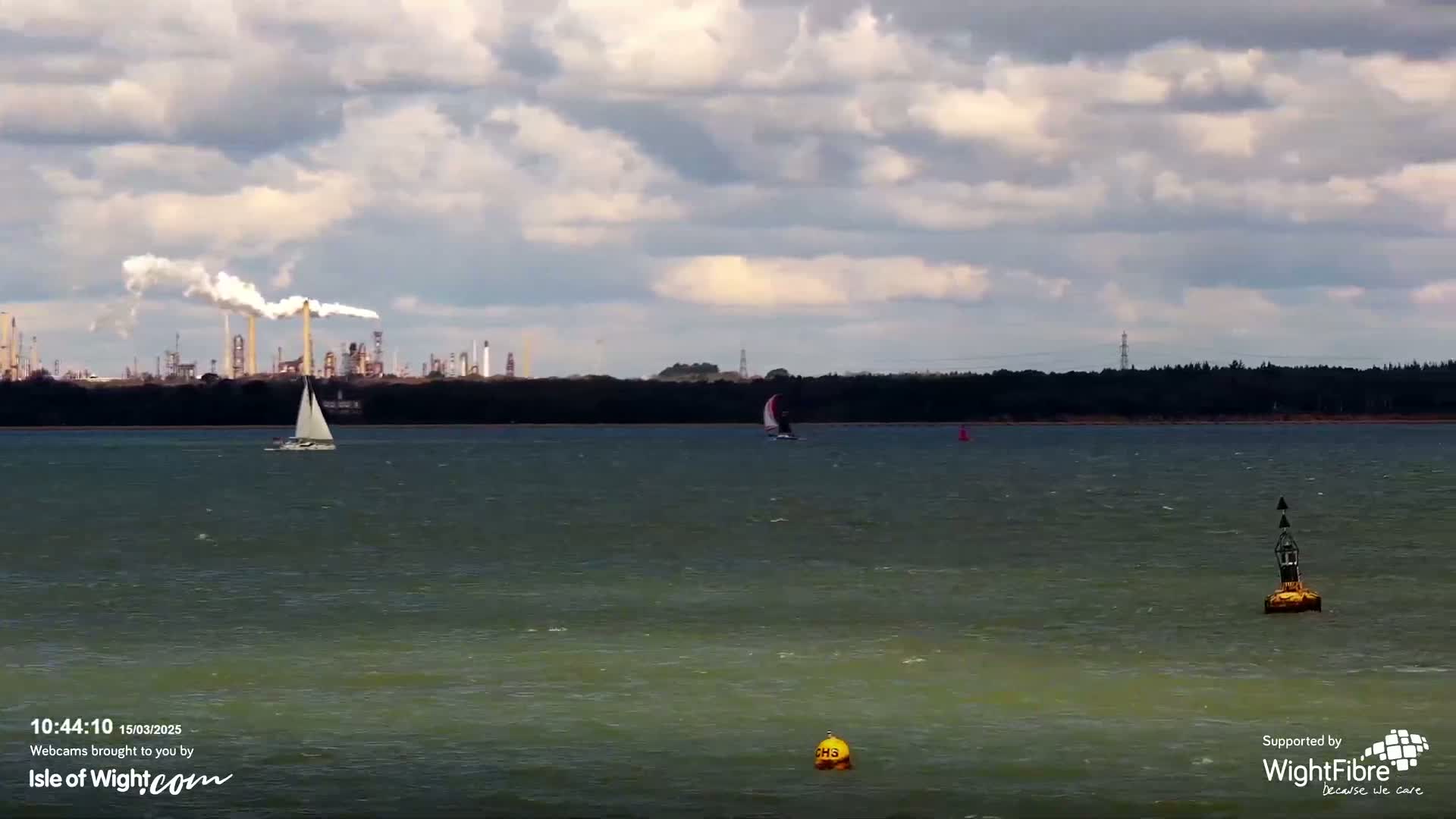 Two sailboats are visible on a choppy body of water under a cloudy sky, with an industrial plant emitting smoke in the distance.